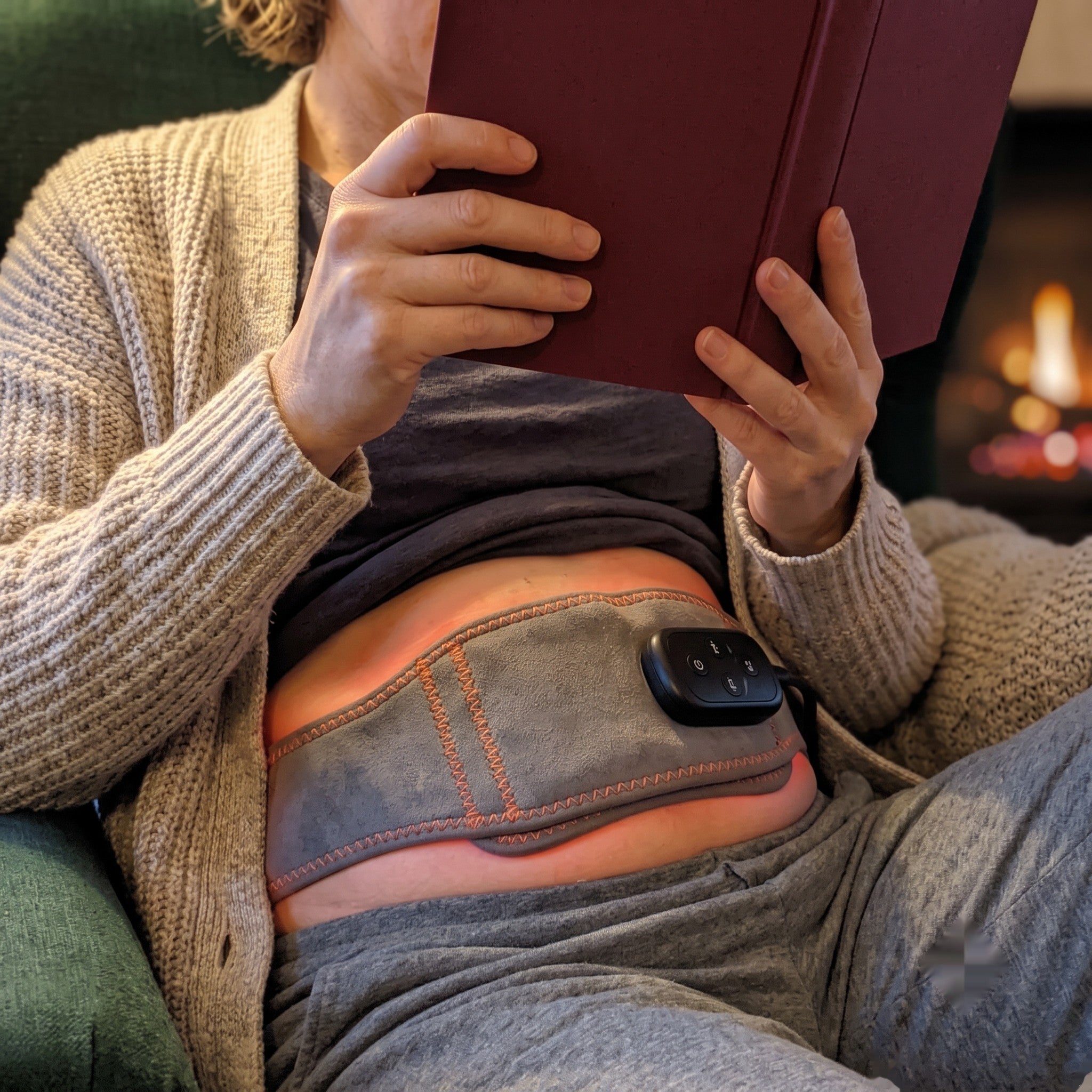 Person relaxing in a reading chair at dusk with the belt visible under an open cardigan, a faint red glow and a book in hand
